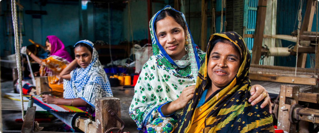 Two women's smile for the camera while other in the background continue to work at a clothing mill.